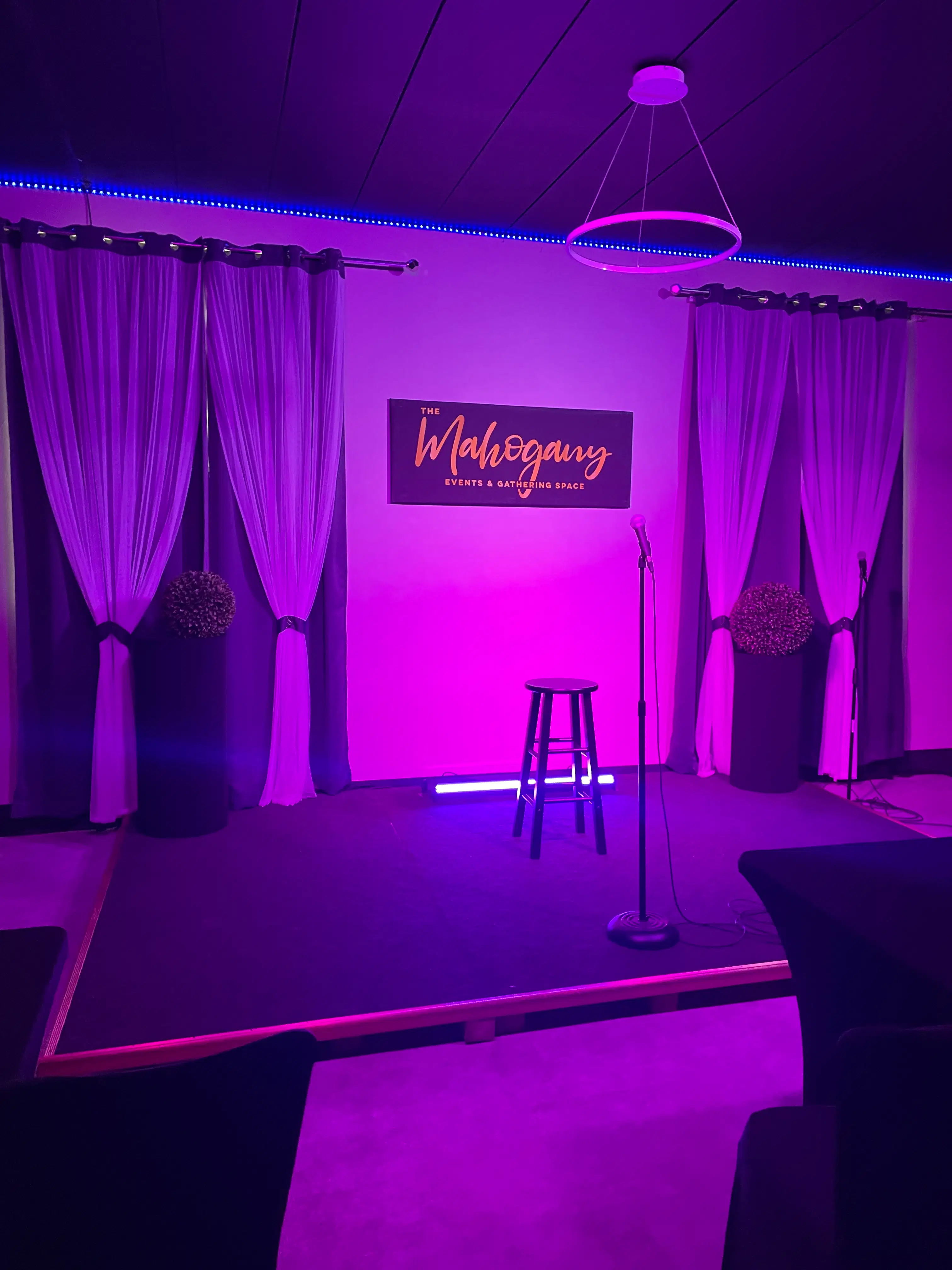 A microphone stand and stool on The Mahogany's stage under purple lighting